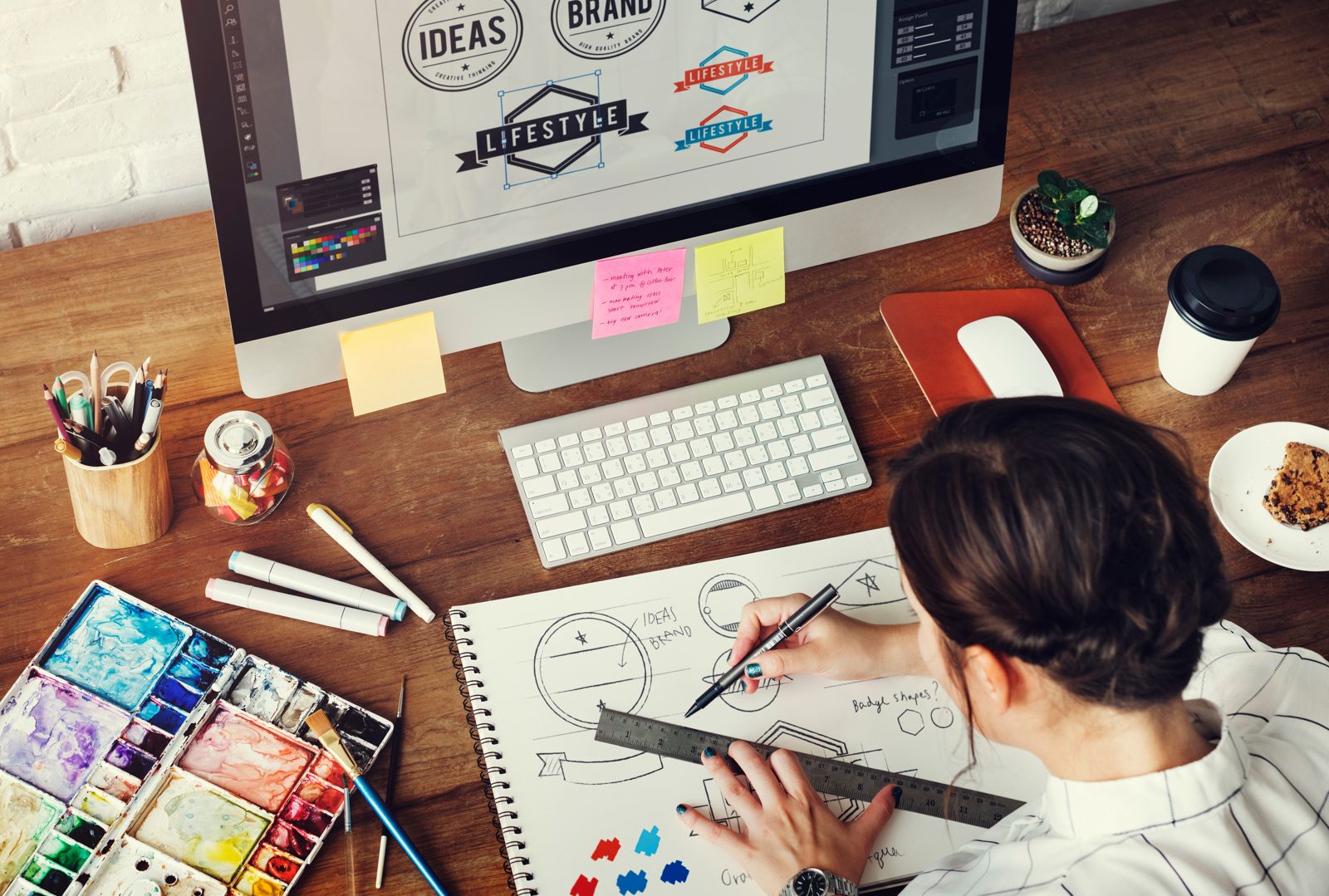 A designer sketching concepts in a notebook beside the design on screen