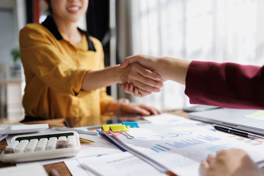 Two people shaking hands across a desk covered in performance reports